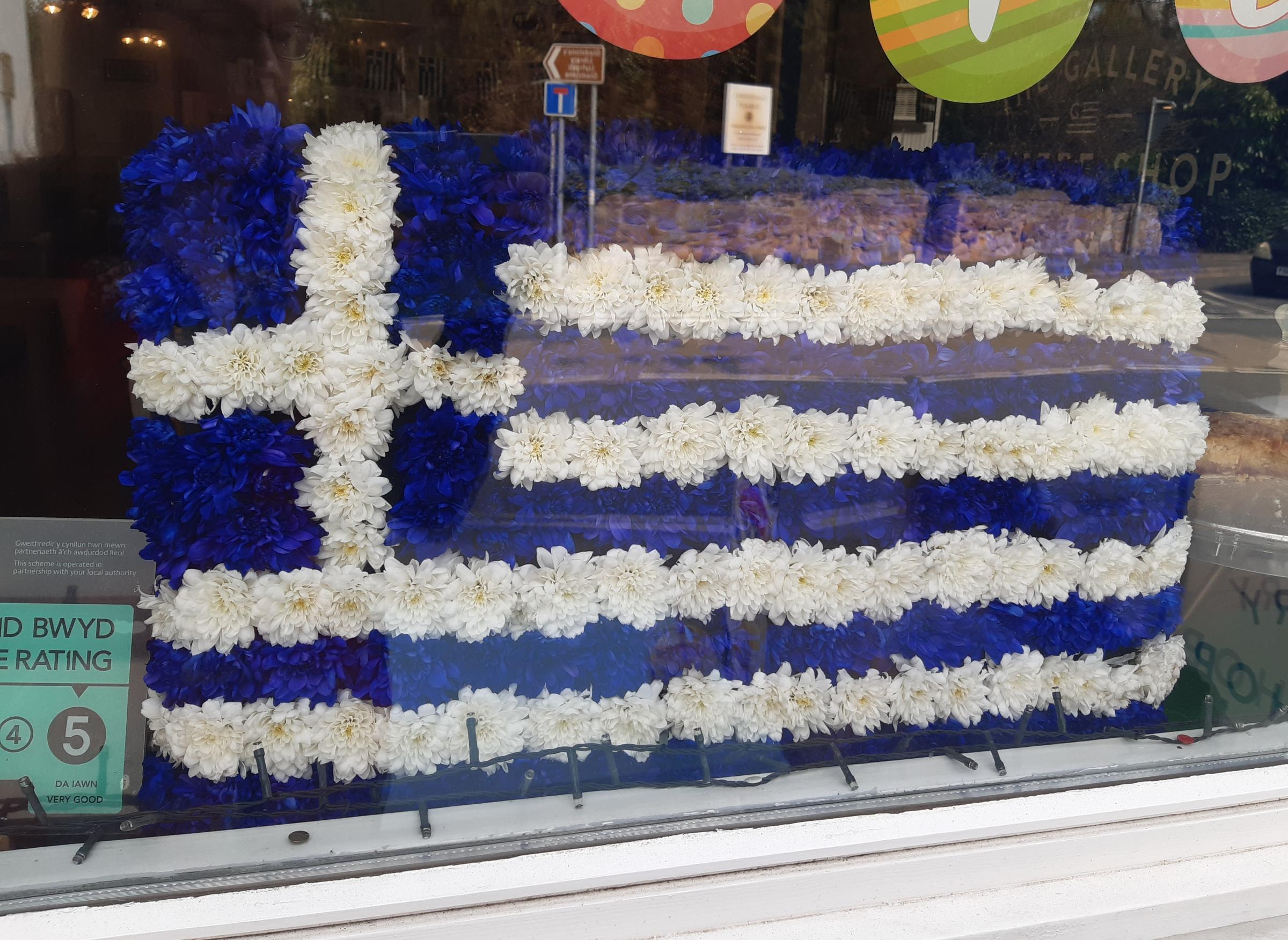 Greek flag floral tribute to Spiros Kavvadias, in the window of The Gallery, in Hawarden.